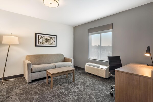 A modern hotel room with a grey sofa, a wooden coffee table, a desk with a lamp, a standing lamp, and a large window with a city view.