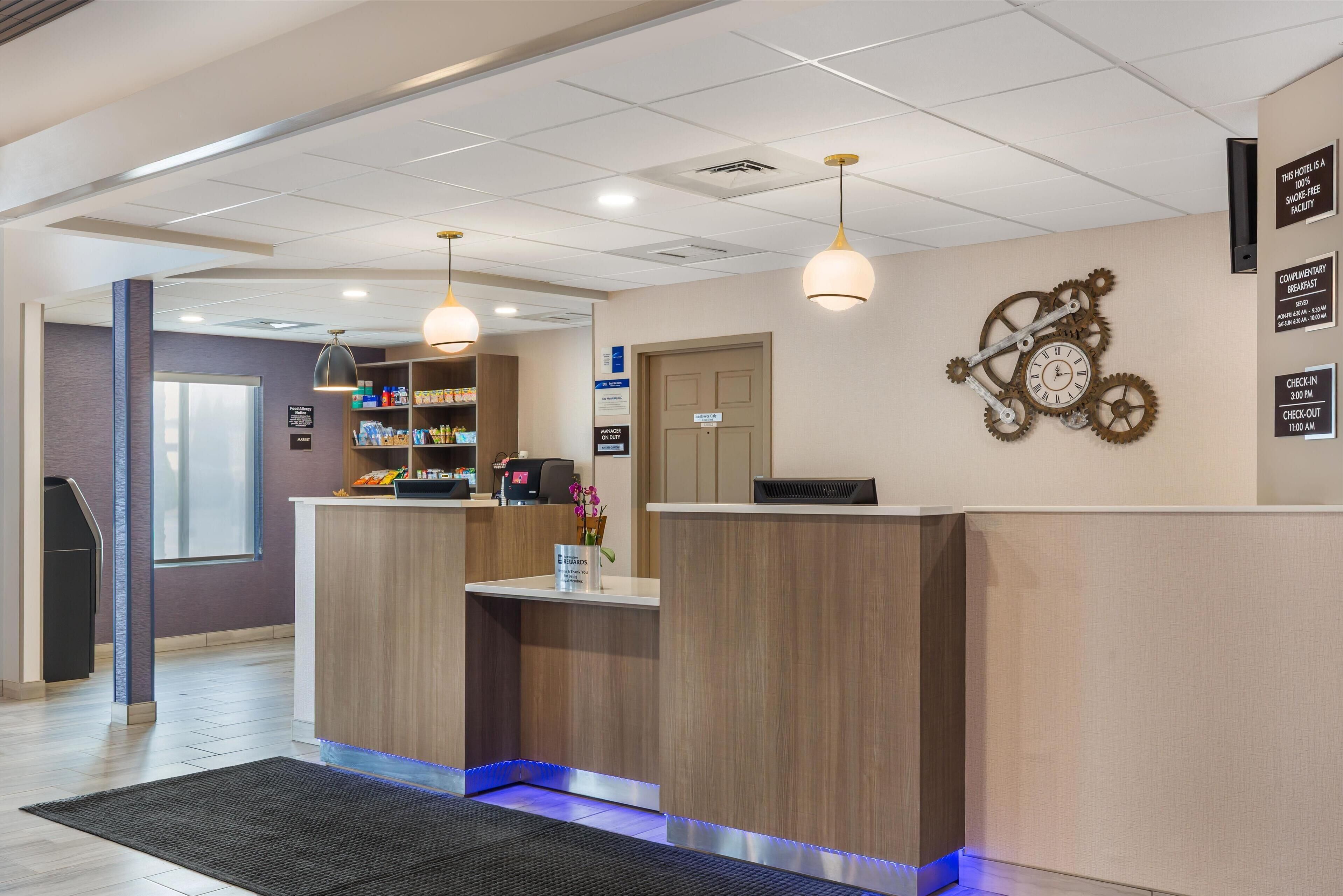 A hotel reception area with a wooden reception desk, a clock with gears on the wall, and a sign indicating check-in and check-out times.