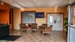 Un hall de hotel con mostrador de recepción, zona de estar con sillas y una mesa, un televisor montado en la pared y un amplio ventanal con cortinas.