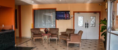 Un hall de hotel con mostrador de recepción, zona de estar con sillas y una mesa, un televisor montado en la pared y un amplio ventanal con cortinas.
