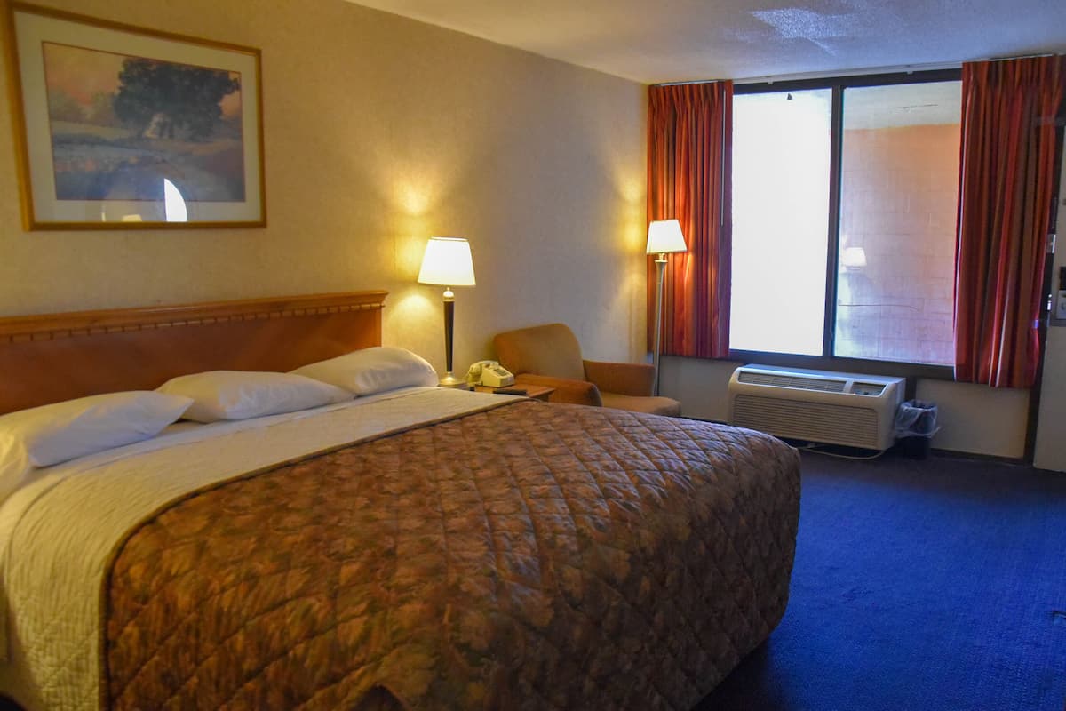 A hotel room with a large bed, two bedside lamps, a chair, a window with curtains, and an air conditioning unit.