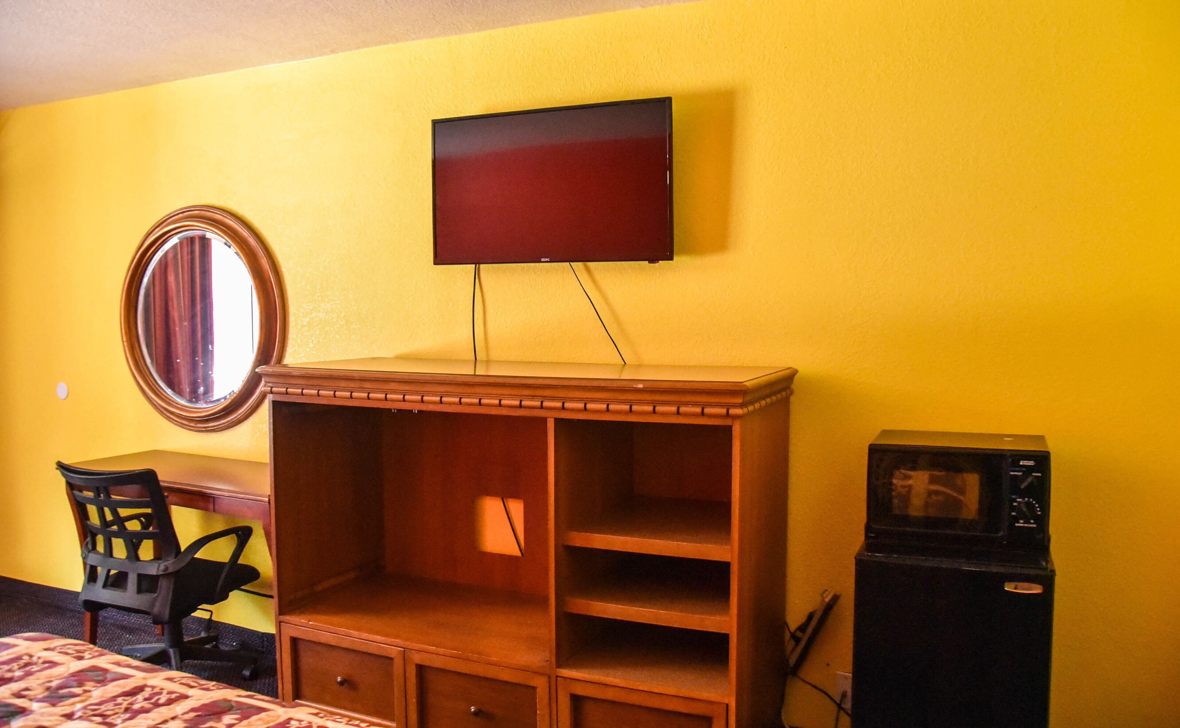 Una habitación con un escritorio de madera, una silla, un televisor montado en la pared y un pequeño electrodoméstico negro.