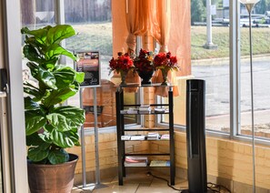 A corner of a room with a large potted plant, a shelf with books, a vase with flowers, and a window with a view of the outside. - Homeplace Inn (Jacksonville)
