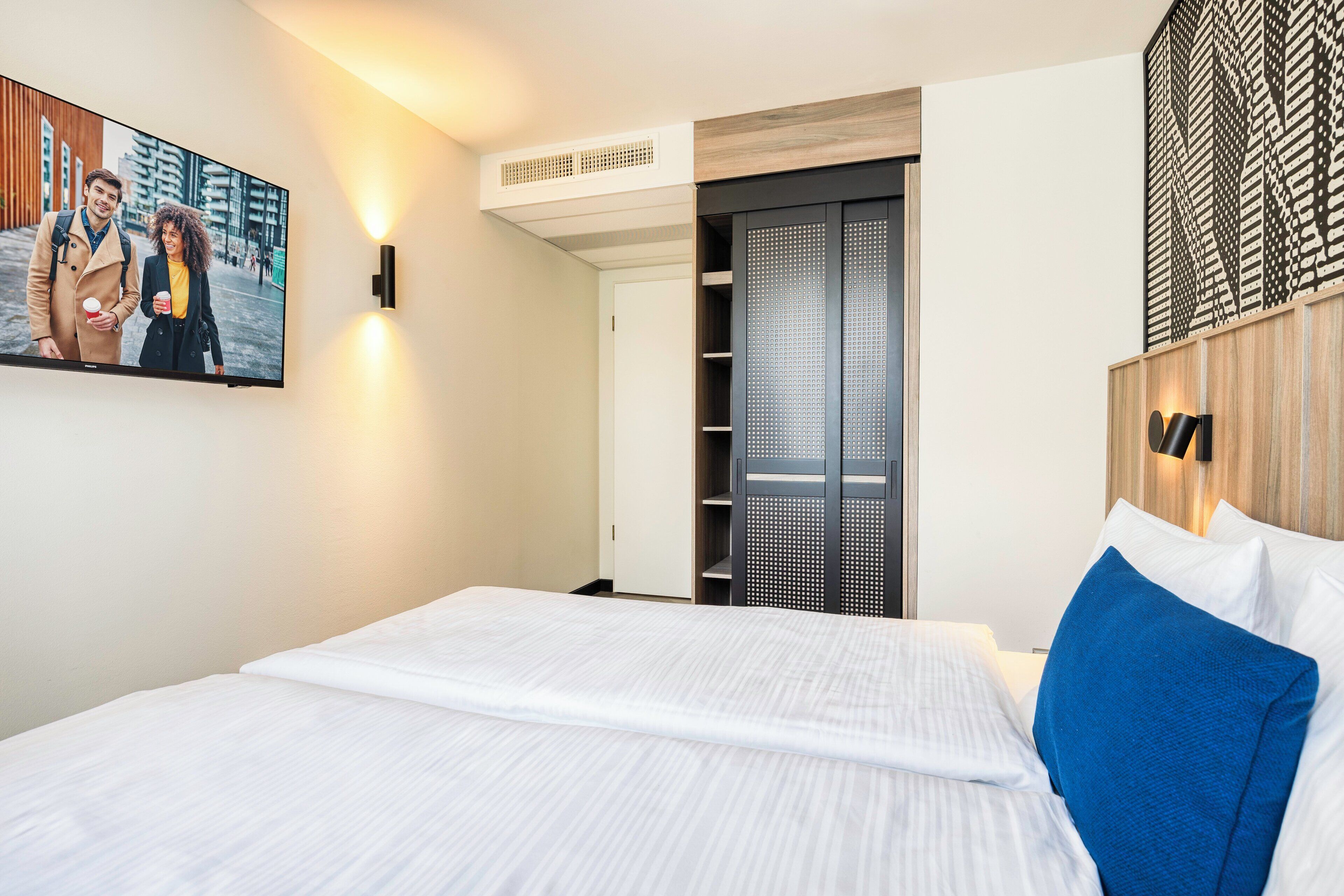Ett modernt hotellrum med en stor säng, en väggmonterad TV, en garderob och en dekorativ panel.