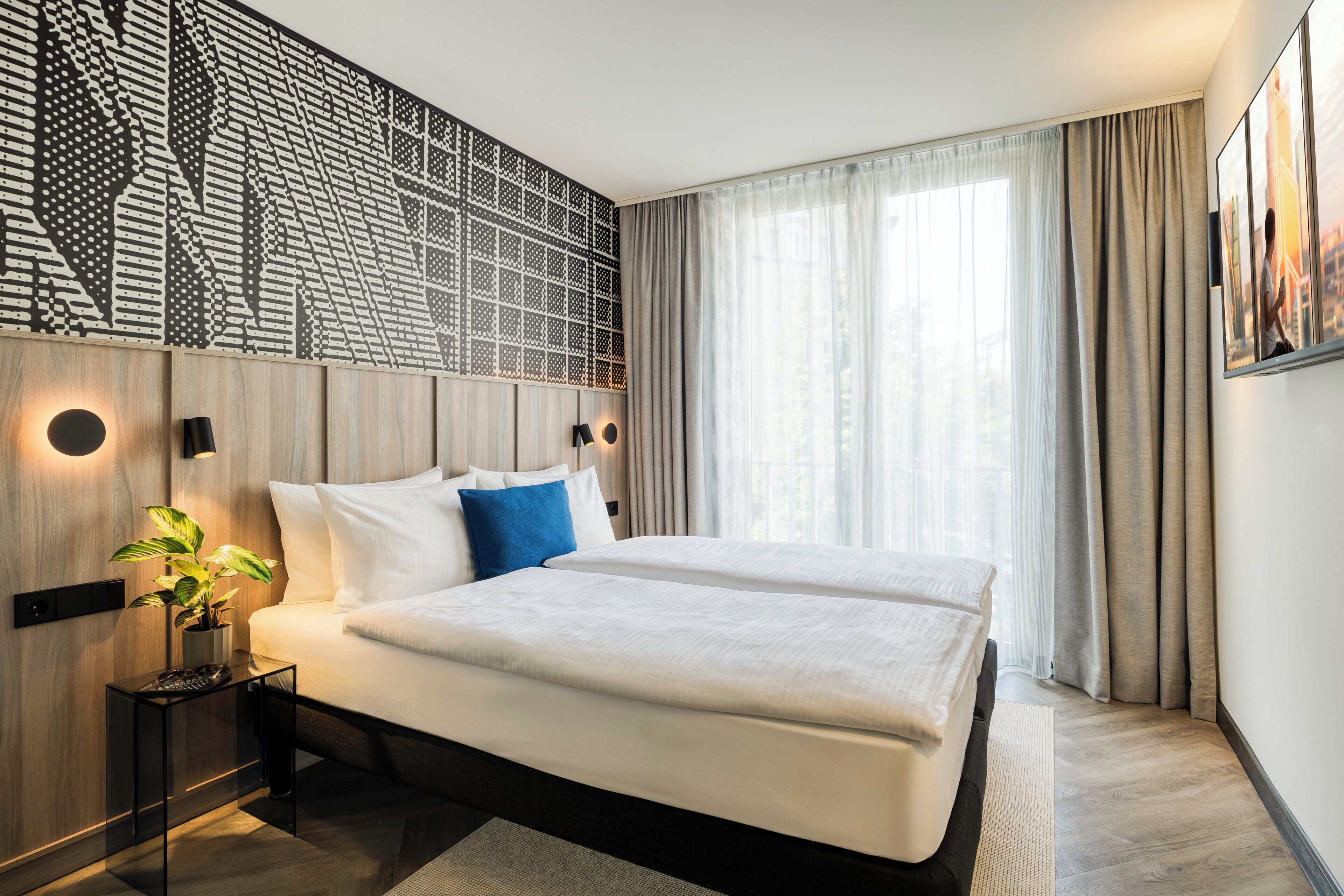 A modern hotel room with a large bed, a decorative wall panel, a bedside table with a lamp, and a large window with curtains.