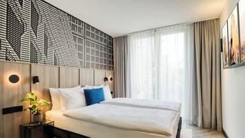 A modern hotel room with a large bed, a decorative wall panel, a bedside table with a lamp, and a large window with curtains.