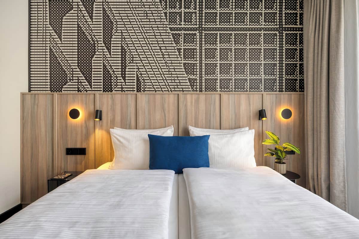 A hotel room with two beds, a wooden headboard, wall-mounted lamps, and a decorative wall panel.