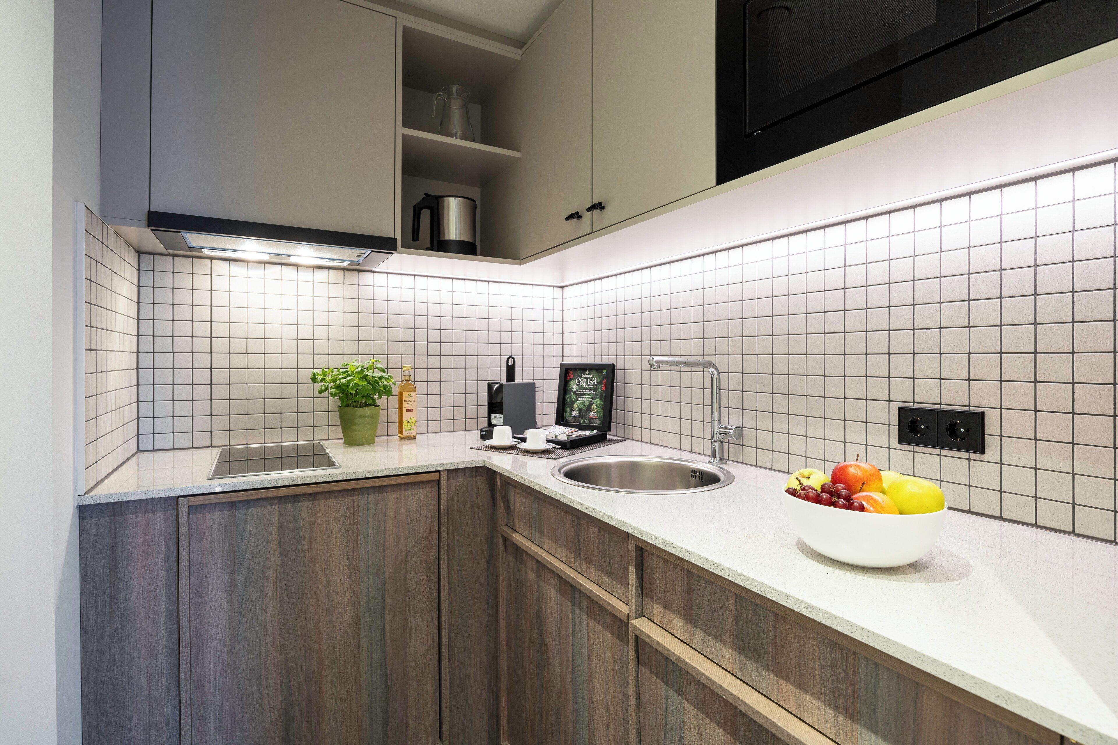 A modern kitchen with white tiles, a stainless steel sink, wooden cabinets, and a bowl of fruit on the counter.
