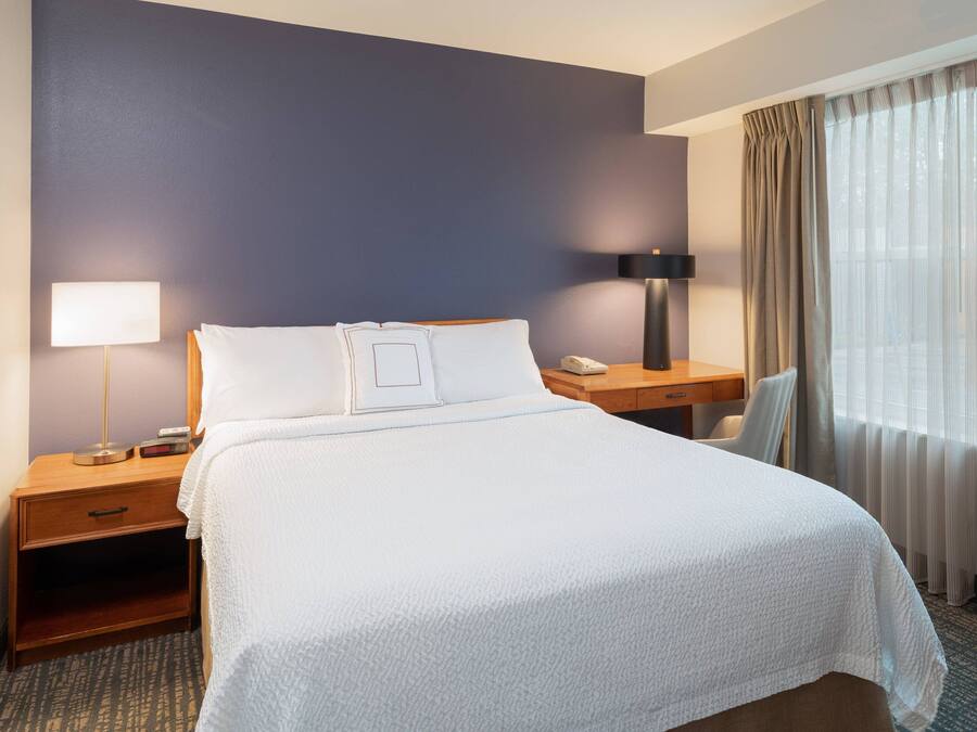 A hotel room with a large bed, a bedside table with a lamp, a desk, and a chair.