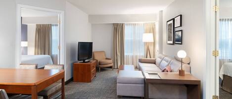 Habitación de hotel con mesa de comedor de madera, sofá, televisor y escritorio con lámpara.