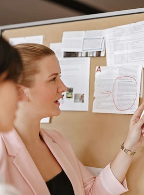 A person in a pink blazer pointing at a bulletin board with papers and a red circled note.