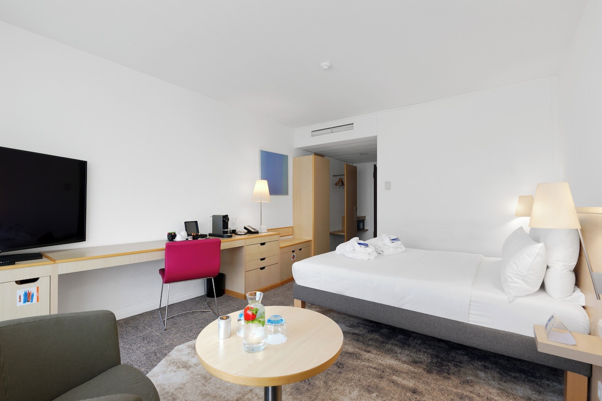 Una habitación de hotel moderna con cama, escritorio, silla, televisor y una pequeña mesa redonda con una botella y vasos.