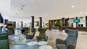 A spacious lobby with modern seating, a reception desk, and a large screen displaying information. - Novotel Berlin Mitte (Berlin)