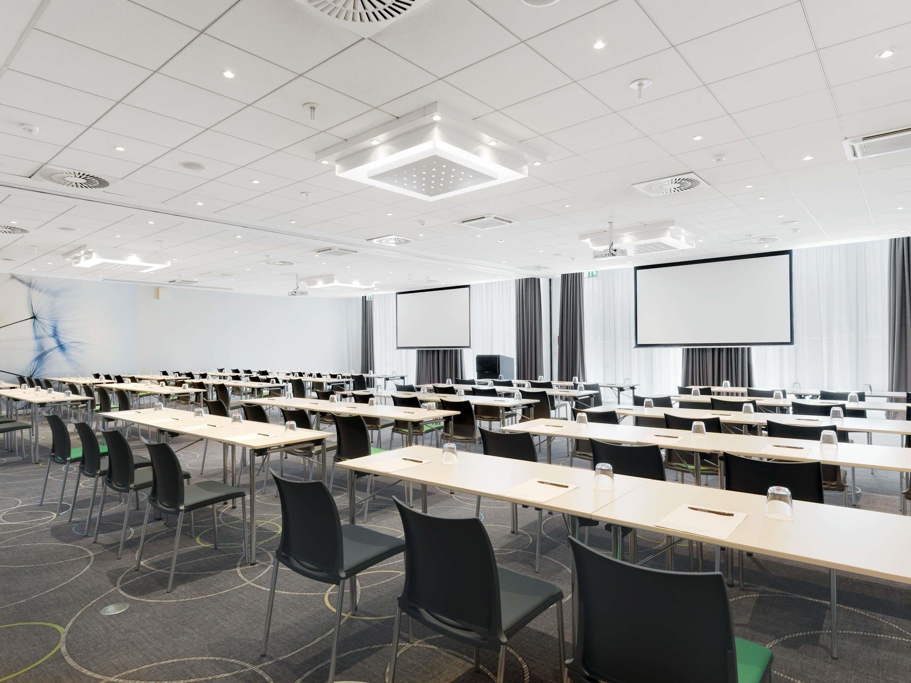 Une salle de conférence équipée de plusieurs tables, de chaises, d’un écran de projection et d’un grand tableau blanc.