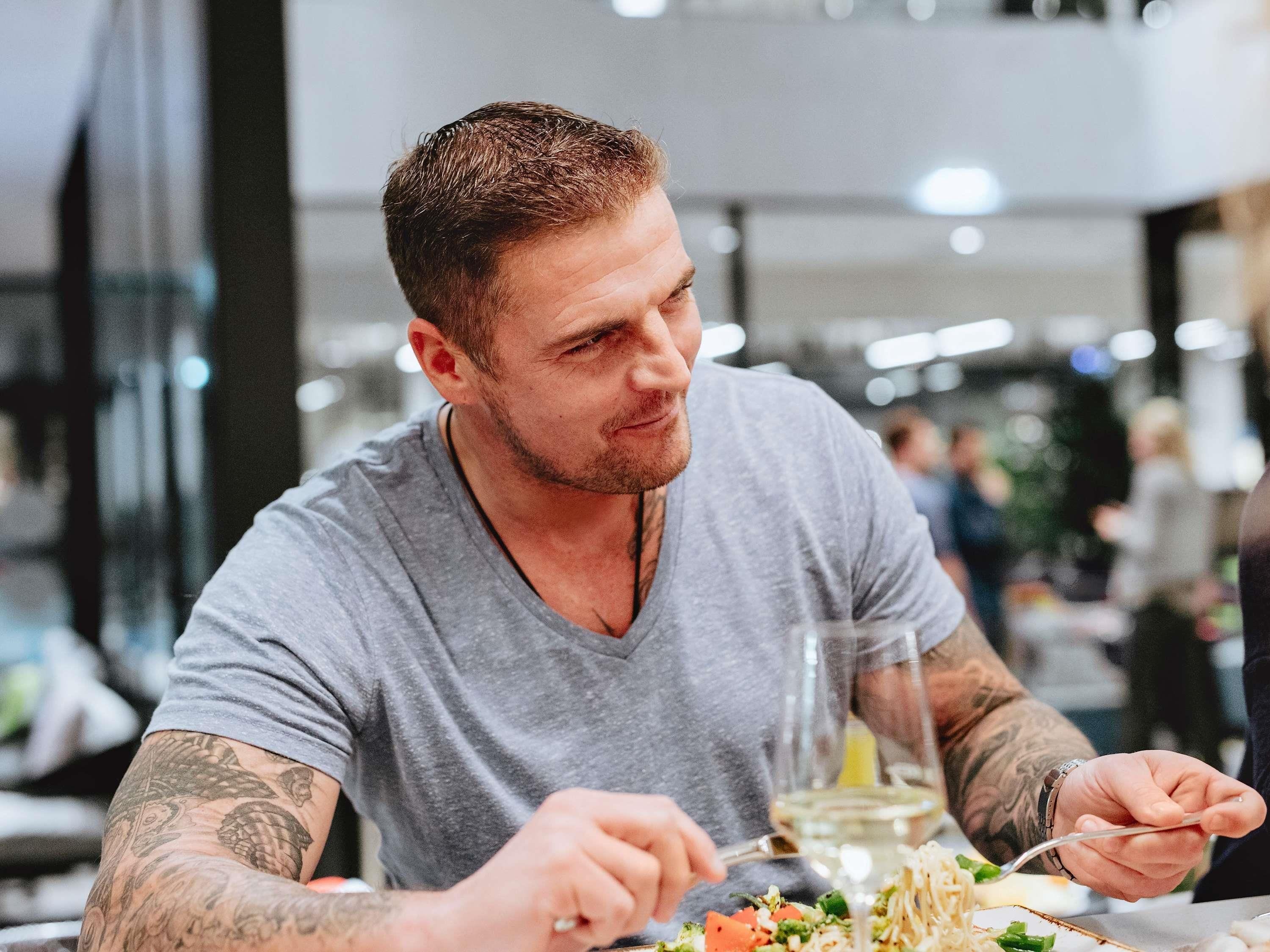 Una persona con tatuajes cenando en un restaurante, con una copa de vino blanco.