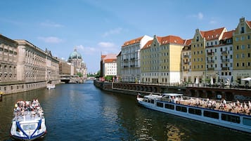 A river with a boat carrying passengers, buildings with balconies, and a prominent dome structure in the background.