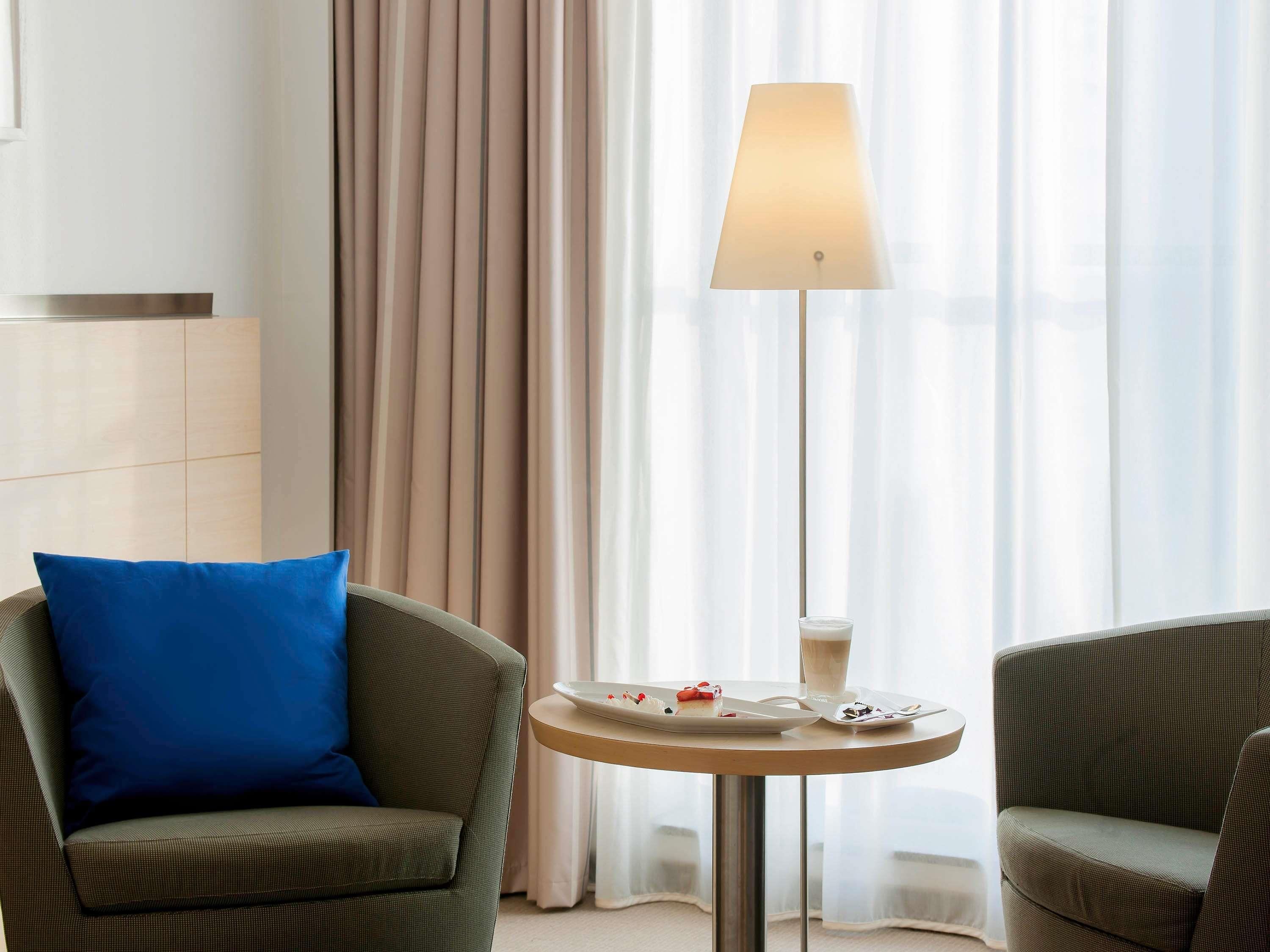 Habitación de hotel con sofá, una pequeña mesa redonda con un plato de comida y un vaso de bebida, y una lámpara de pie.