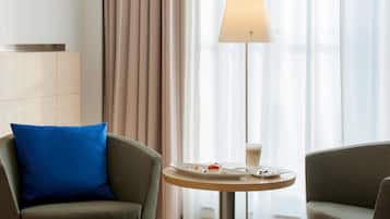 Habitación de hotel con sofá, una pequeña mesa redonda con un plato de comida y un vaso de bebida, y una lámpara de pie.