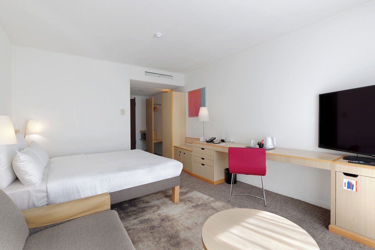 Una habitación de hotel moderna con una cama, un escritorio, una silla, un televisor y un armario.