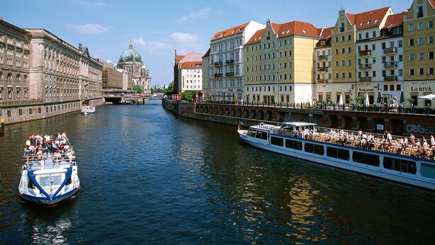 A river with a boat carrying passengers, buildings with balconies, and a prominent dome structure in the background. - Novotel Berlin Mitte (Berlin)