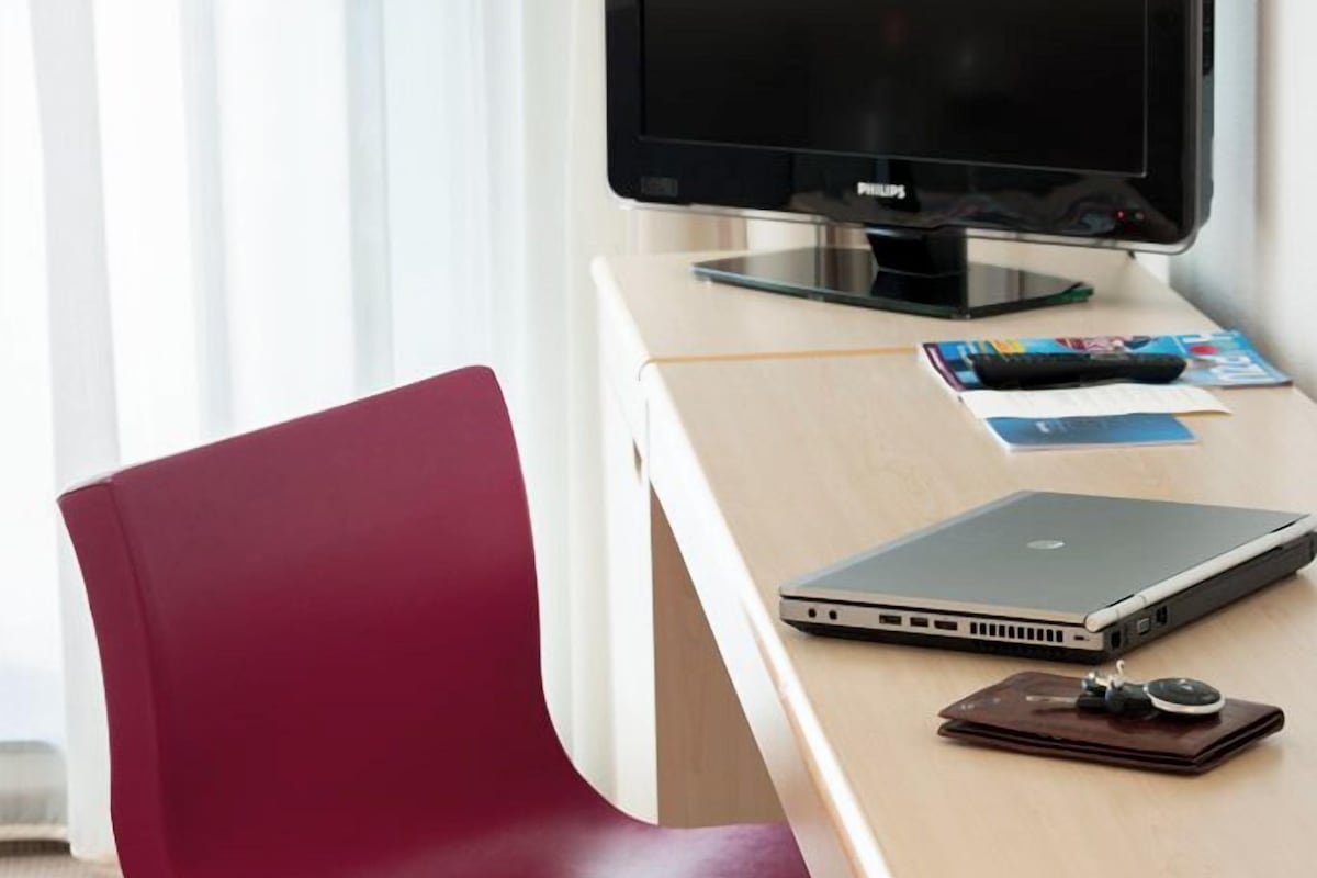 Una habitación de hotel moderna con un escritorio de madera, una silla roja, una computadora portátil, una tableta y un televisor.
