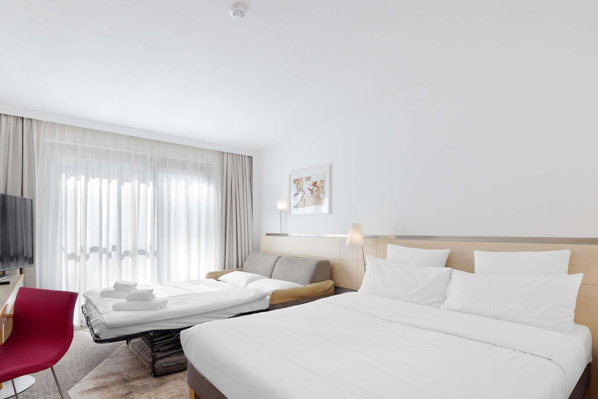 A hotel room with a large bed, a smaller bed, a desk with a chair, a TV, and a window with curtains.