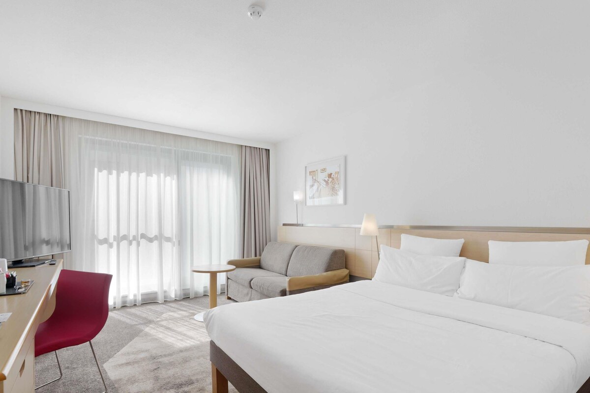 A hotel room with a large bed, a desk with a chair, a TV, and a window with curtains.