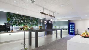 A modern hotel lobby with a central bar, a green wall with 'BERLIN' text, ceiling fans, and a blue-lit beverage dispenser. - Novotel Berlin Mitte (Berlin)