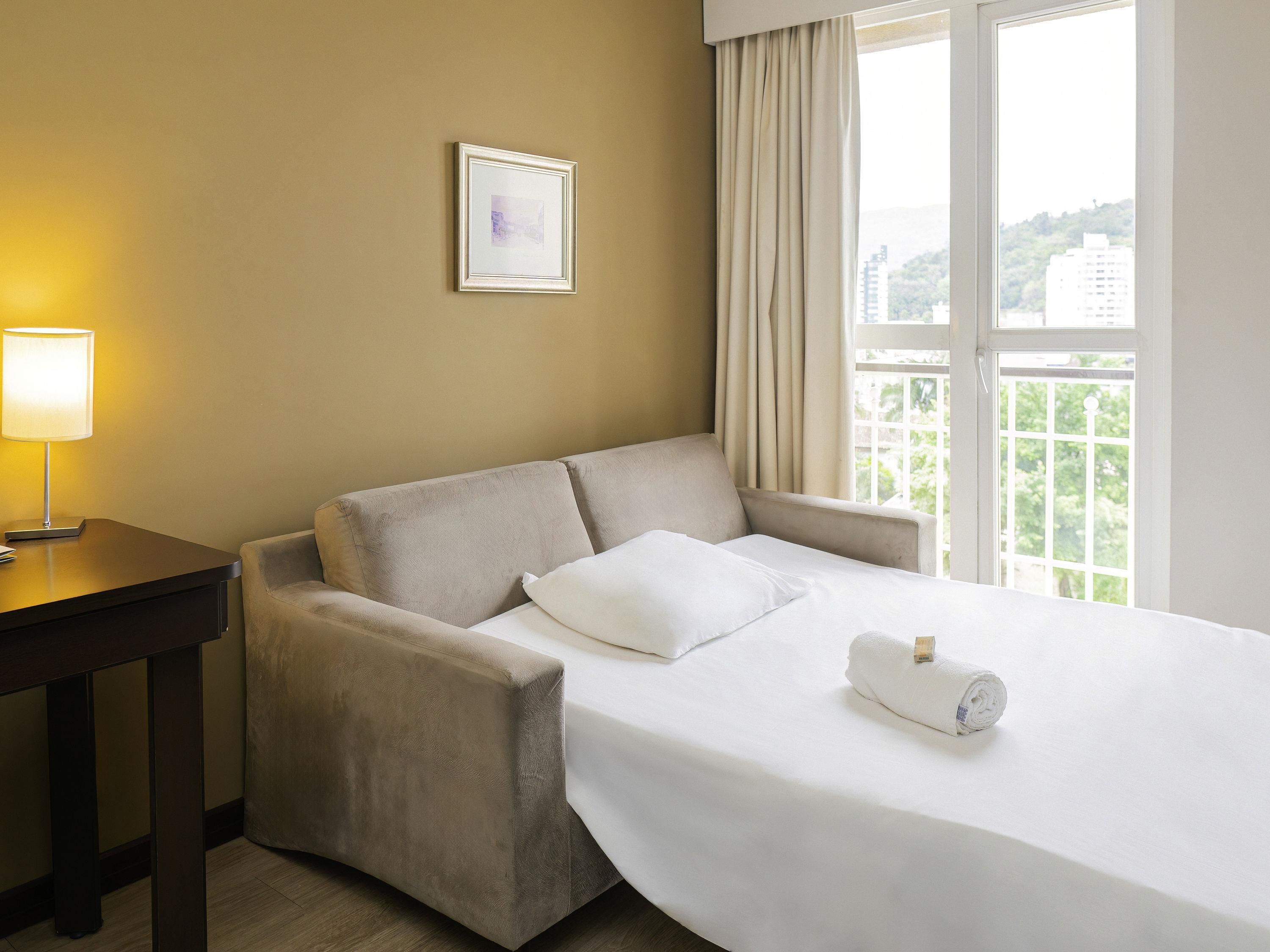 A hotel room with a bed, a chair, a desk, a lamp, and a window with curtains.