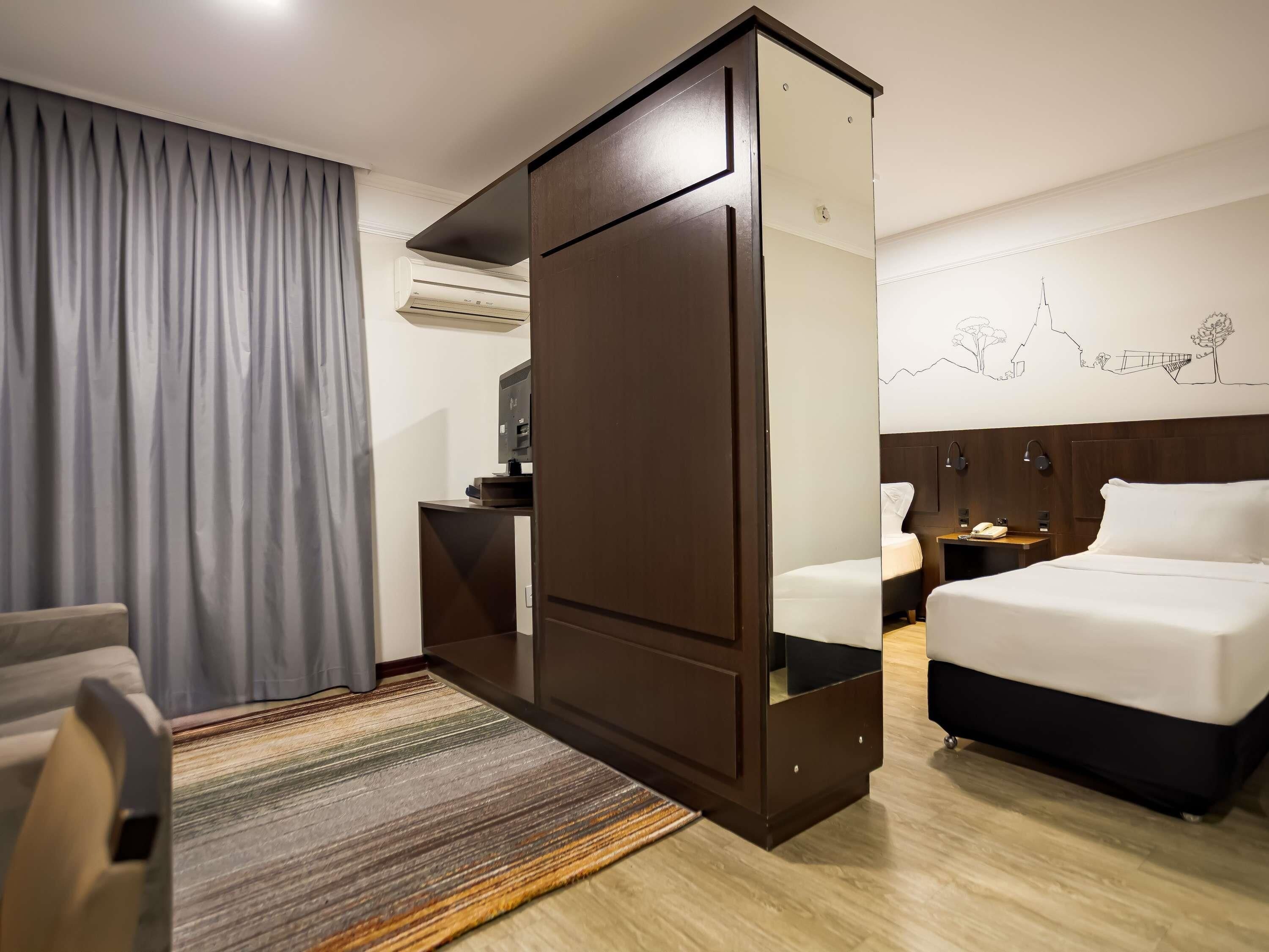 A hotel room with a large bed, a smaller bed, a desk with a computer, a chair, a wardrobe, and a patterned rug.