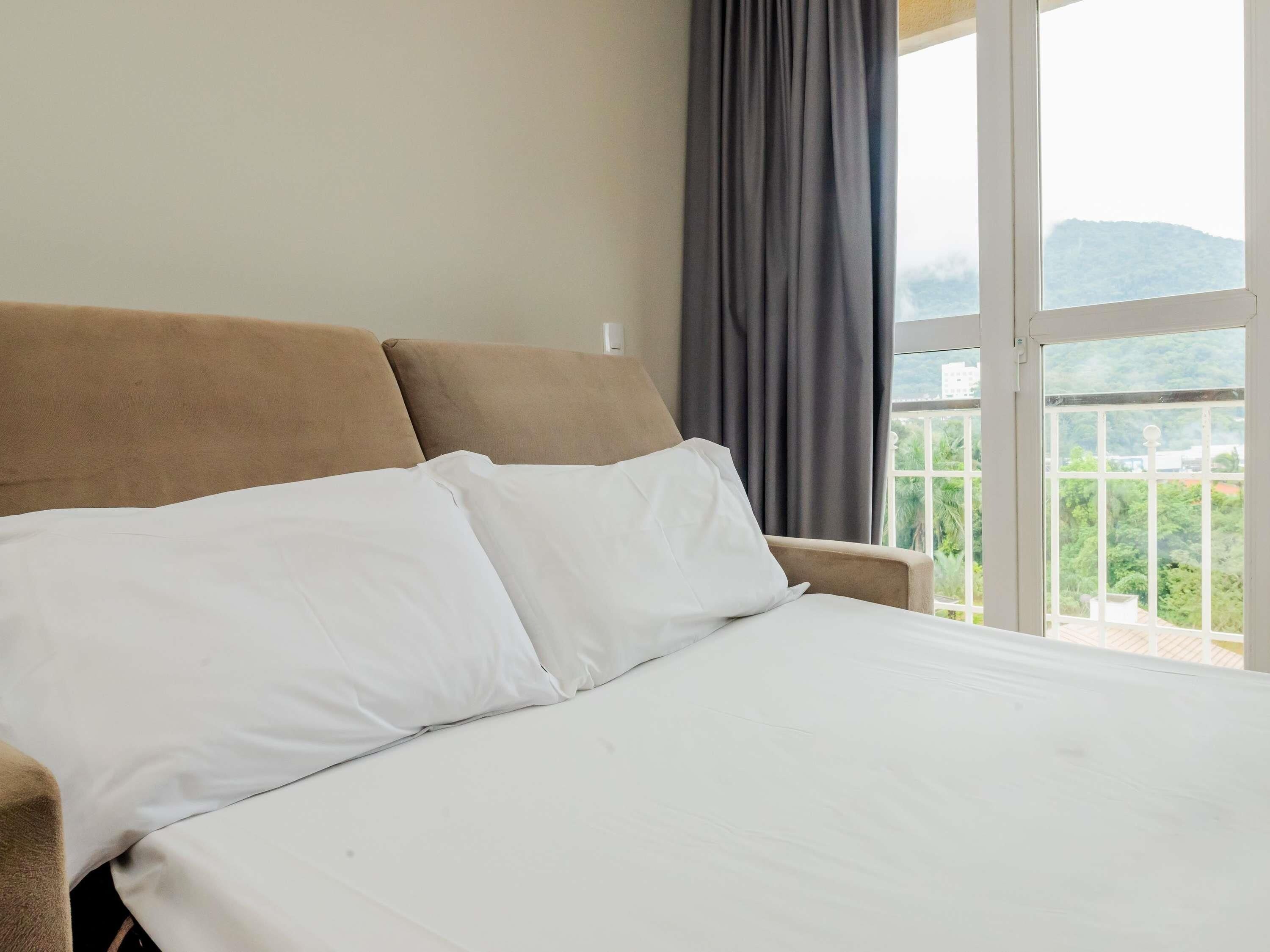 A bed with white linens, a beige headboard, and a view of greenery through a window.