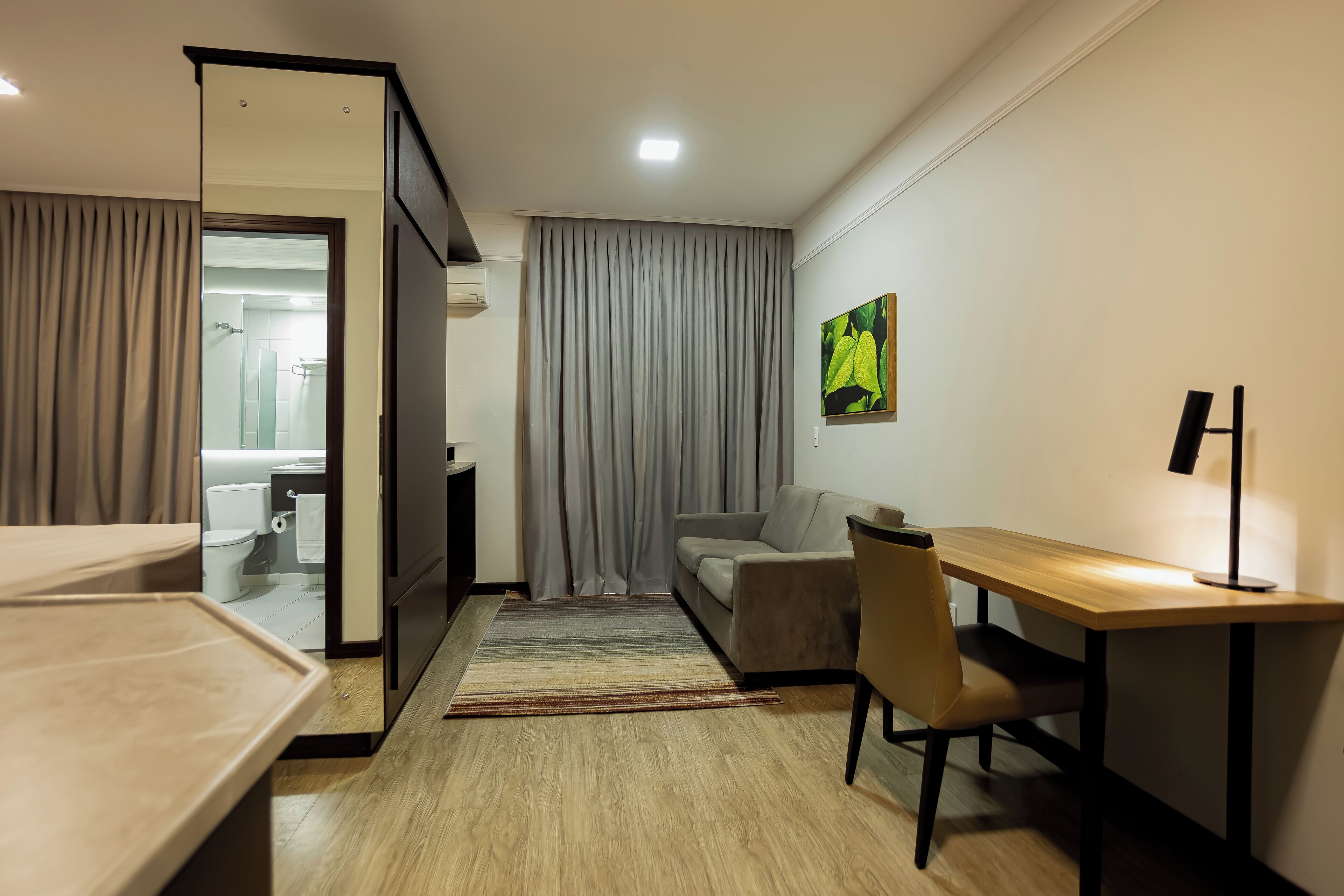 A modern hotel room with a wooden desk, a sofa, a dining table, and a view of the bathroom.