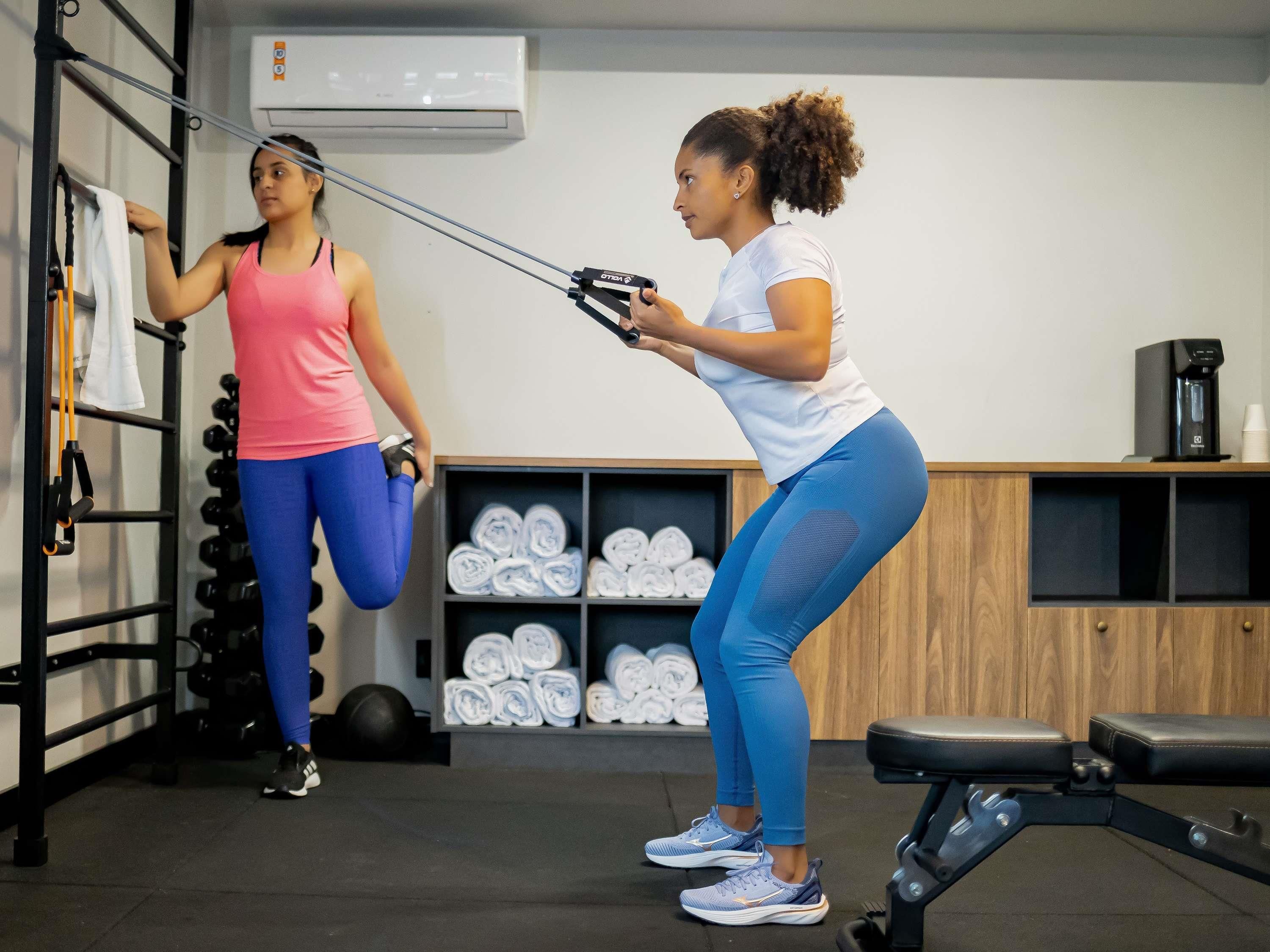 Two individuals exercising with resistance bands in a gym with a rack of dumbbells, a coffee machine, and neatly rolled towels.