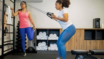 Two individuals exercising with resistance bands in a gym with a rack of dumbbells, a coffee machine, and neatly rolled towels.