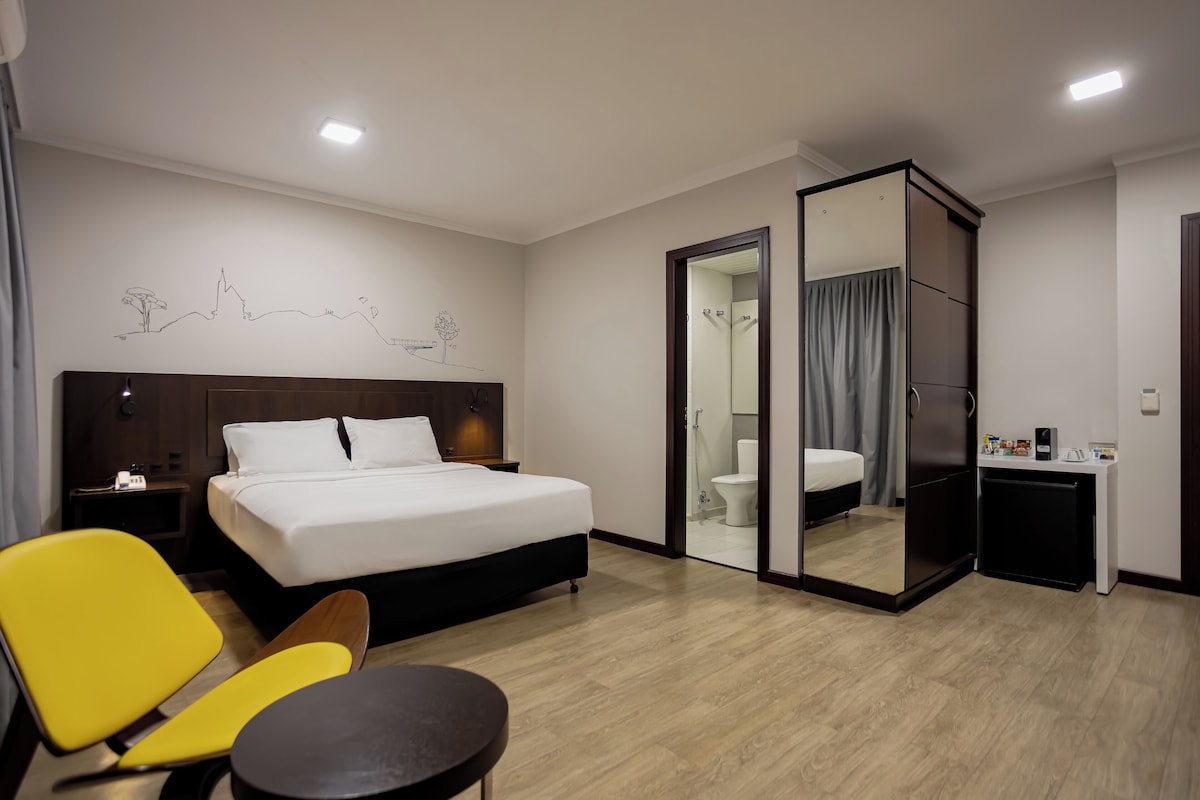 Habitación de hotel con cama, escritorio, silla, espejo y baño visible a través de una puerta de vidrio.