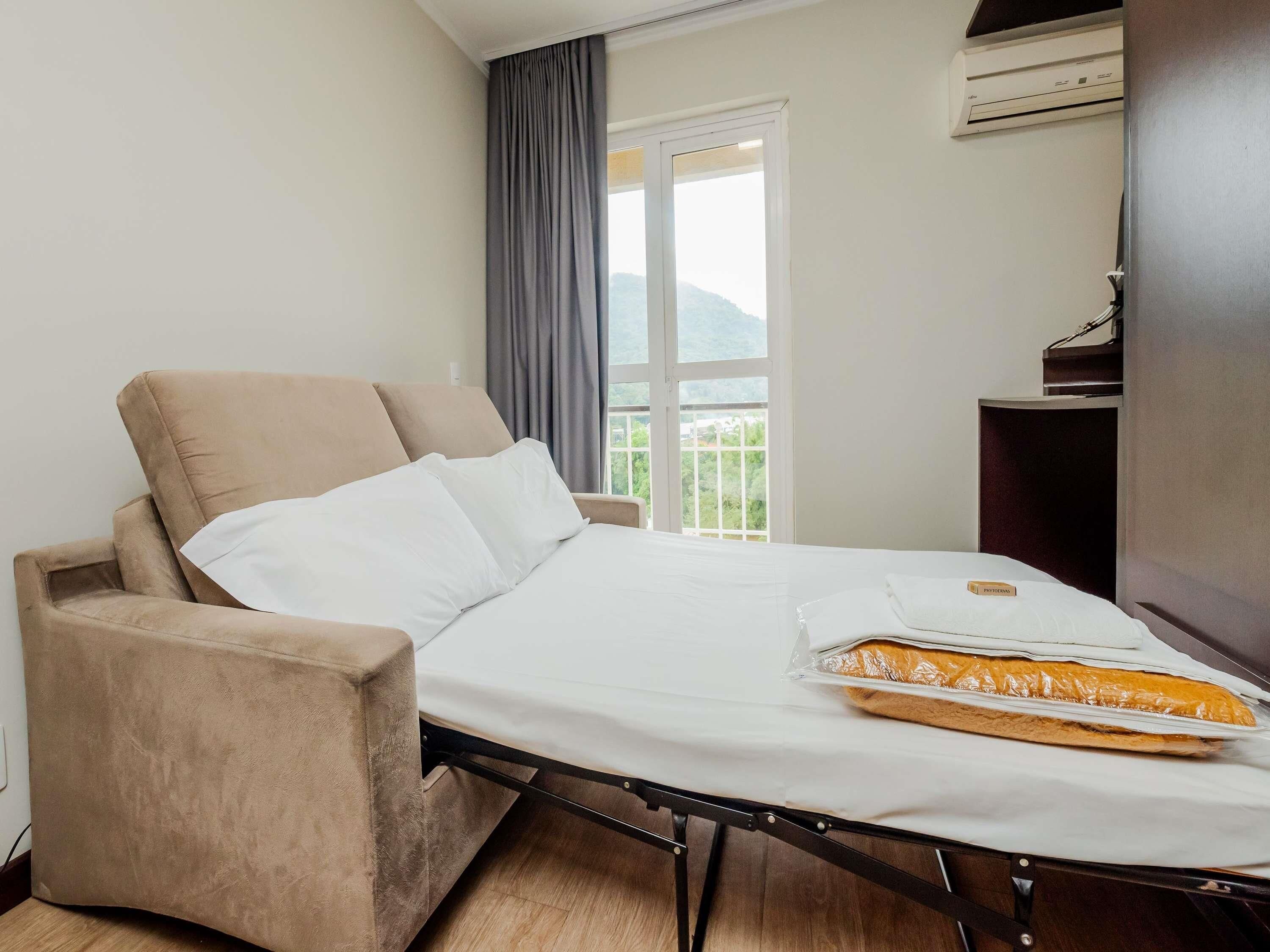 A hotel room with a bed, a chair, a window with a view of greenery, and a small table with a book.