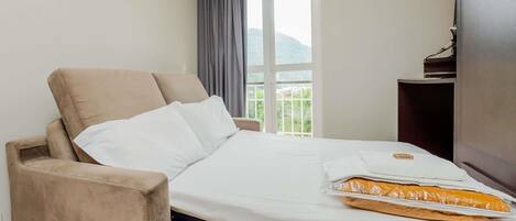 A hotel room with a bed, a chair, a window with a view of greenery, and a small table with a book.