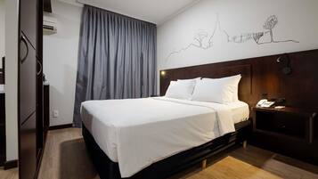 A hotel room with a bed, a nightstand, a phone, a wall-mounted lamp, and a window with curtains.
