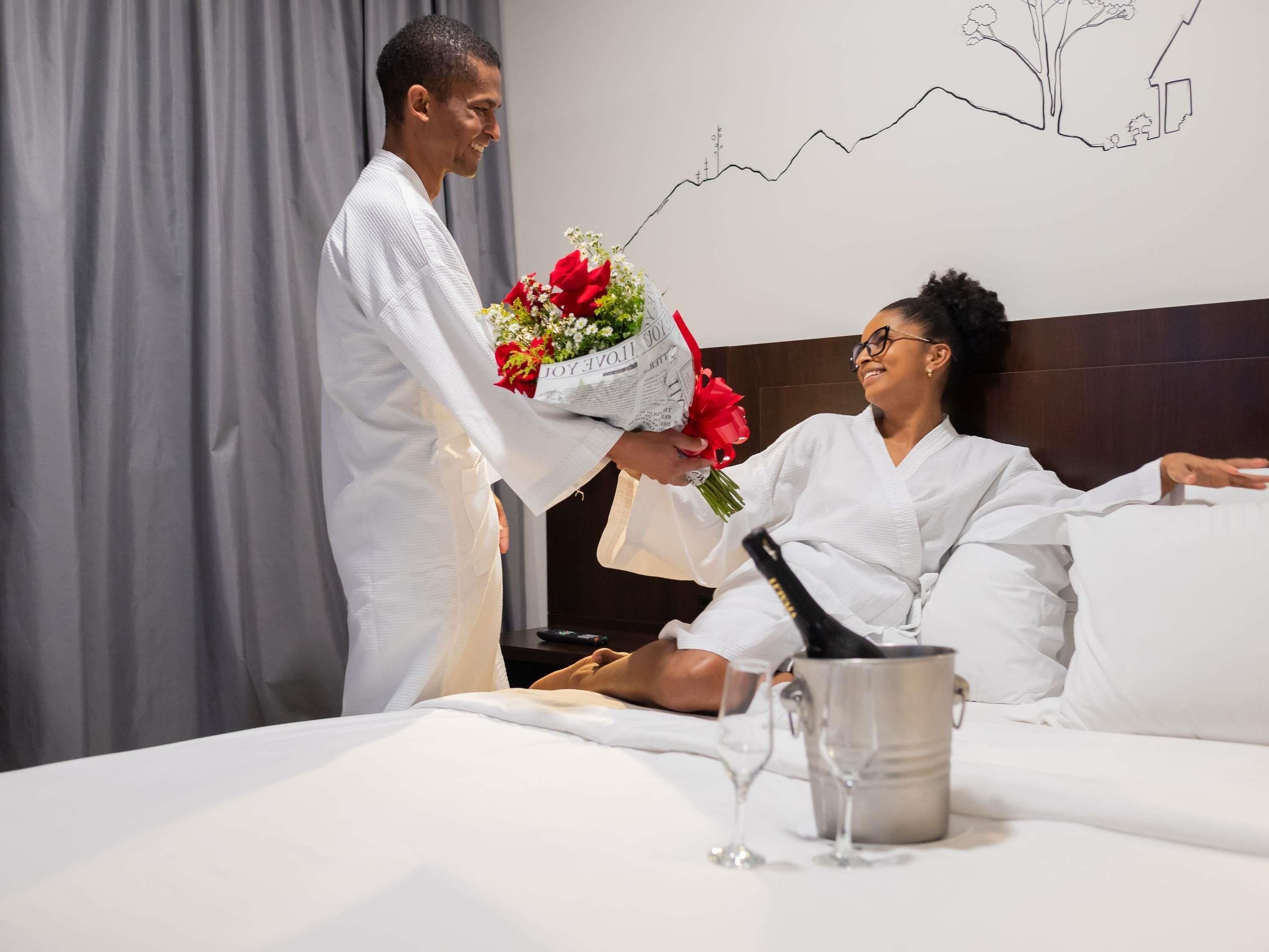 Une personne vêtue d’une robe blanche présente un bouquet de fleurs et une bouteille à une autre personne, également vêtue d’une robe blanche, qui est allongée sur un lit. On aperçoit un verre à vin et un récipient en métal sur le lit.