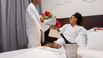 Une personne vêtue d’une robe blanche présente un bouquet de fleurs et une bouteille à une autre personne, également vêtue d’une robe blanche, qui est allongée sur un lit. On aperçoit un verre à vin et un récipient en métal sur le lit.