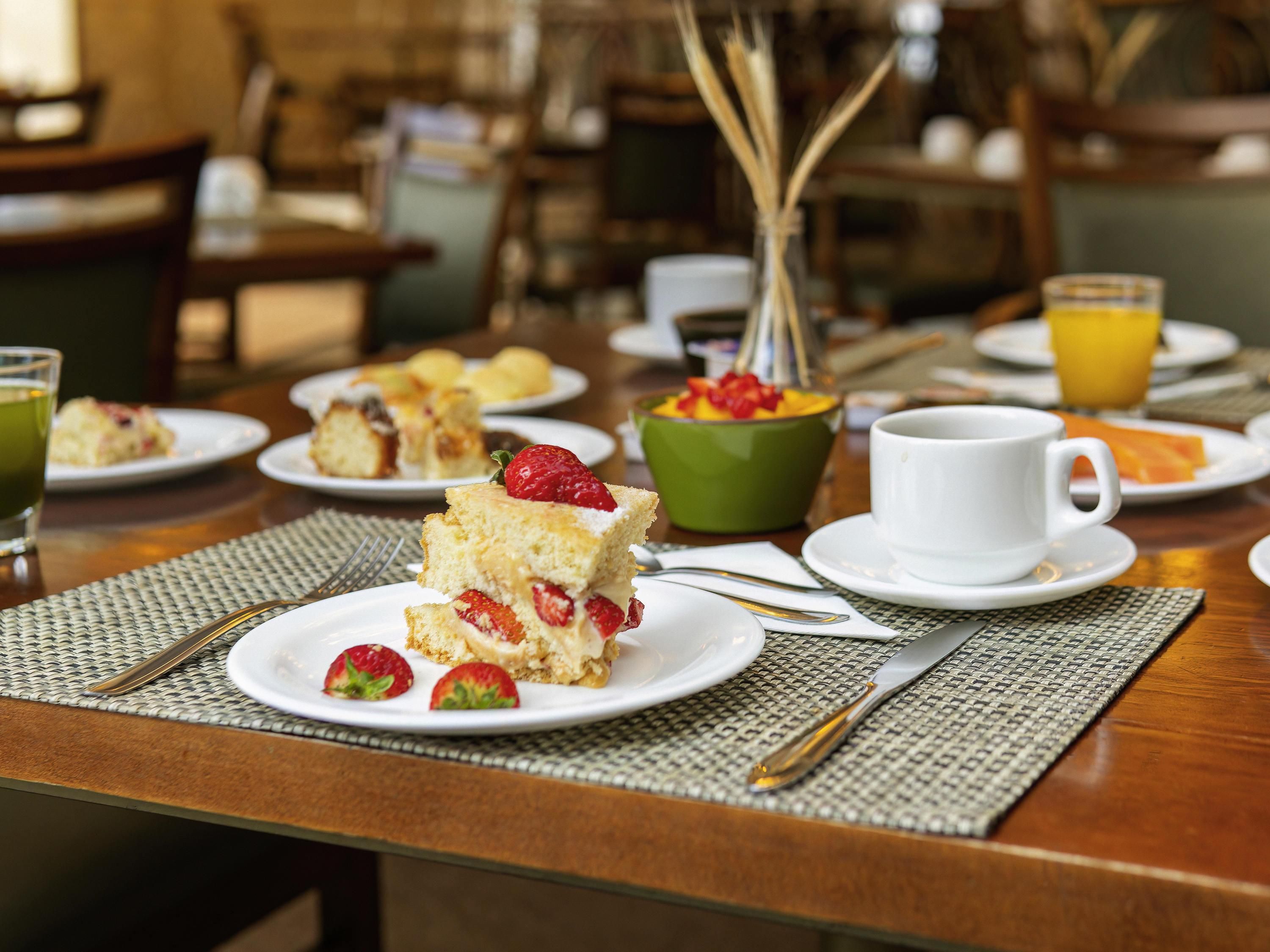 A breakfast setting with a plate of cake, strawberries, a green bowl, a white cup, and a glass of orange juice.