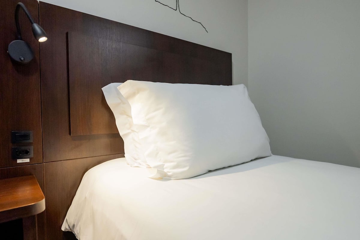 A neatly made bed with white bedding, a wooden headboard, a bedside table with a lamp, and a wall socket.