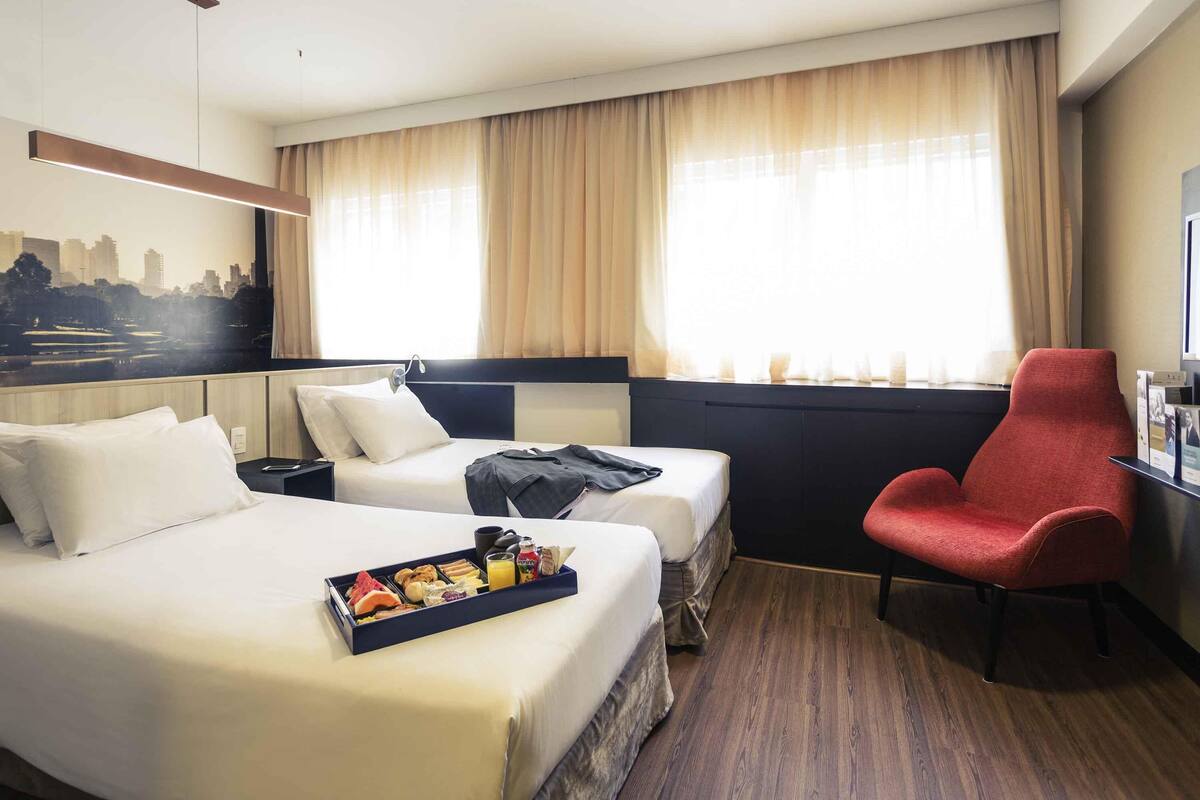 A hotel room with two beds, a red armchair, a cityscape view, and a breakfast tray on the left bed.