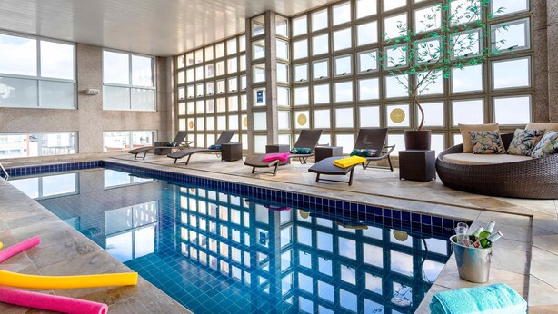 An indoor swimming pool with clear glass walls, multiple lounge chairs, and a seating area with cushions. - Mercure Sao Paulo Bela Vista (São Paulo)