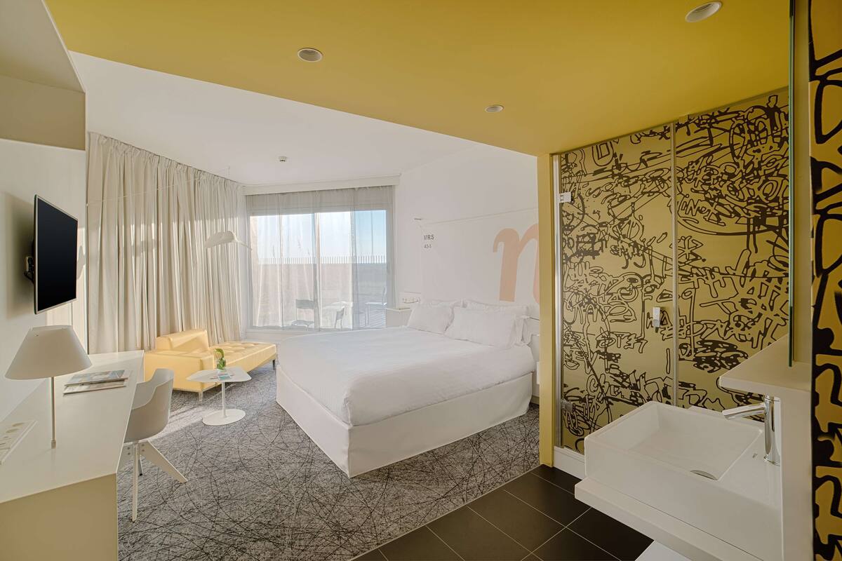 Una habitación de hotel moderna con una cama grande, un escritorio, una silla, una mesita, un televisor y un baño con lavamanos y espejo.