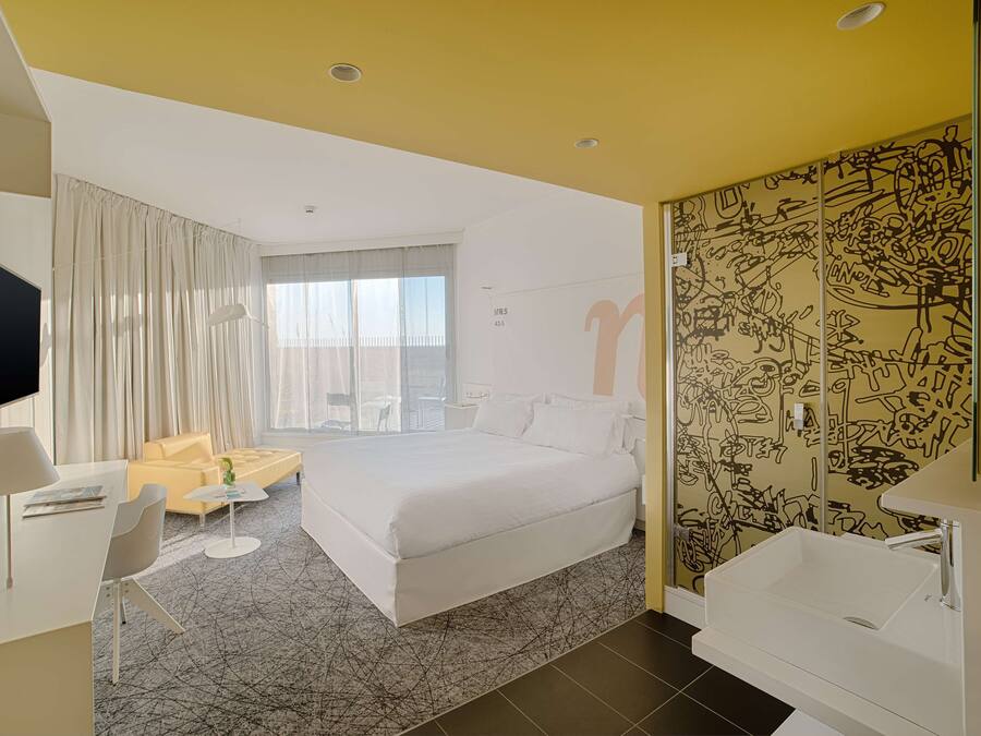Een moderne hotelkamer met een groot bed, een bureau, een stoel, een tafeltje, een televisie en een badkamer met een wastafel en spiegel.