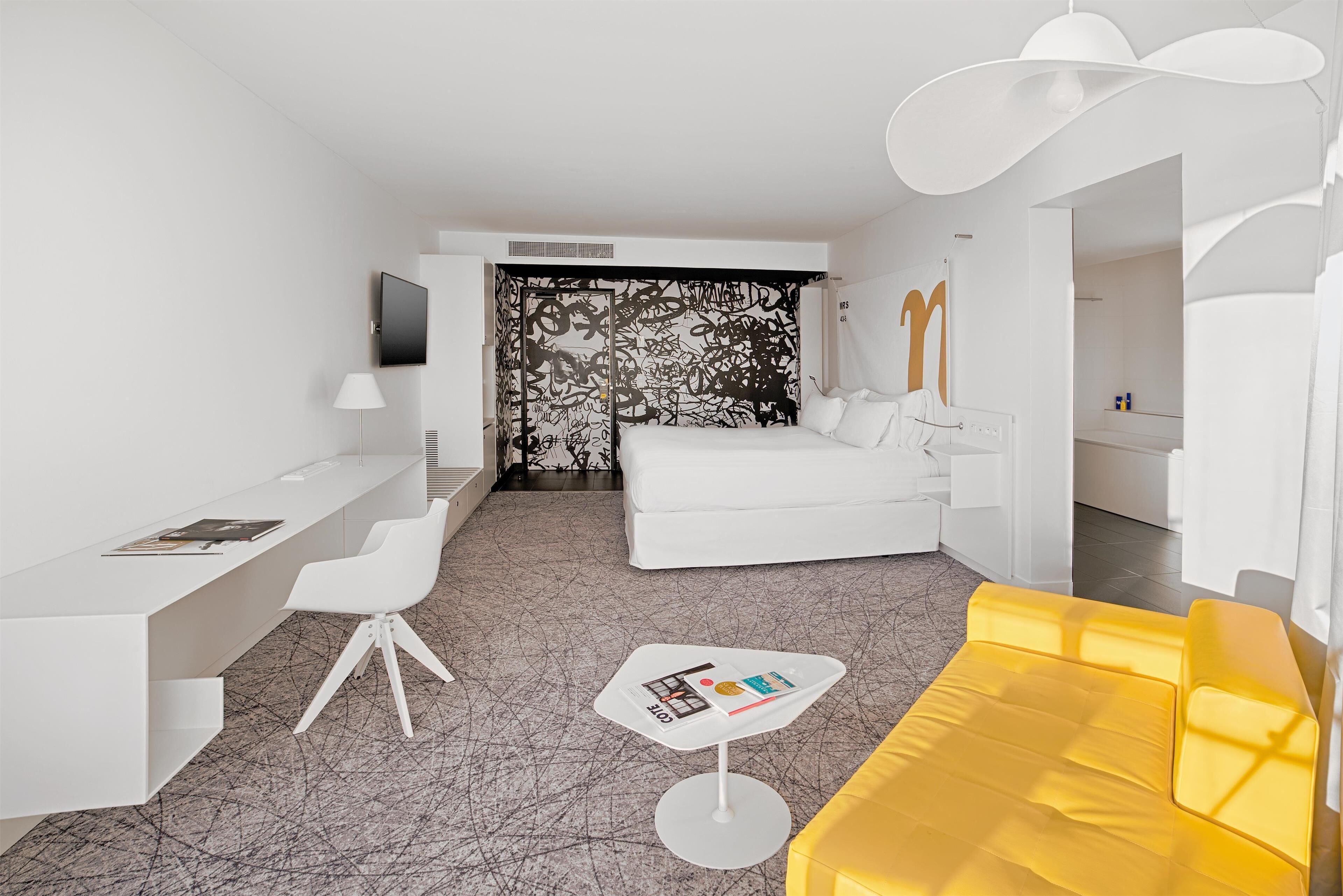 A modern hotel room with a white desk, a yellow sofa, a white armchair, and a patterned curtain.