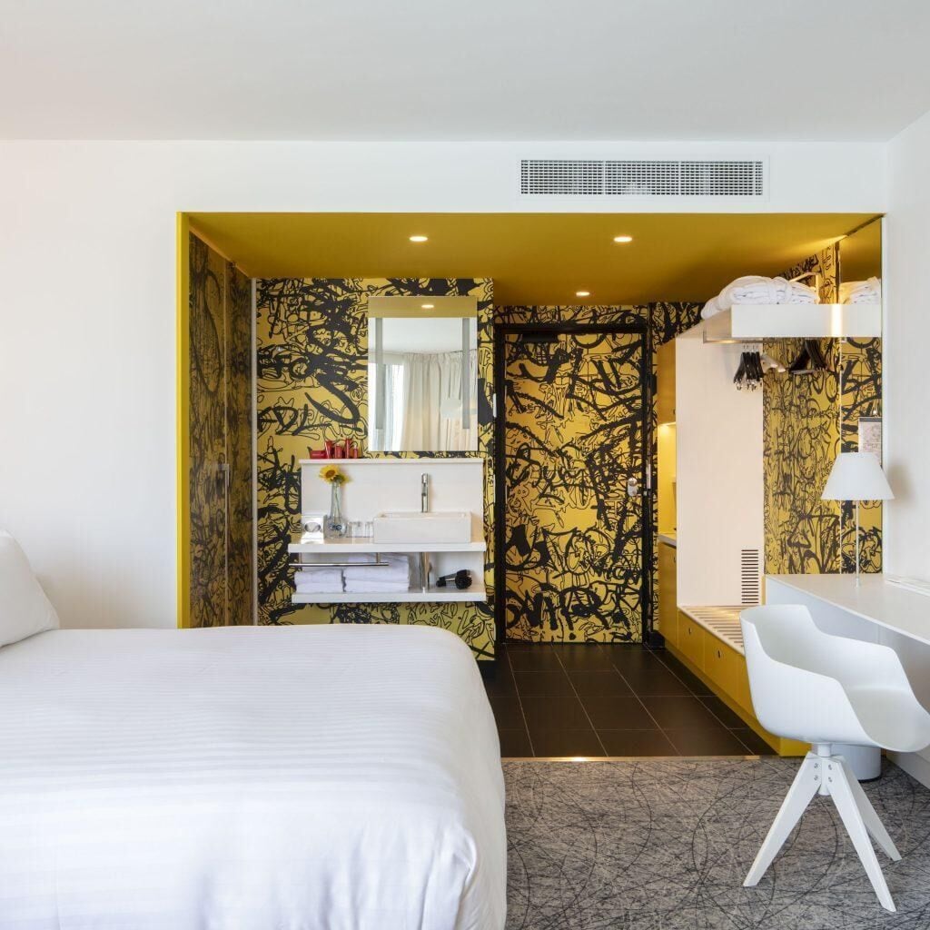 Habitación de hotel con cama, escritorio, baño con lavabo y espejo, y pared con diseño.