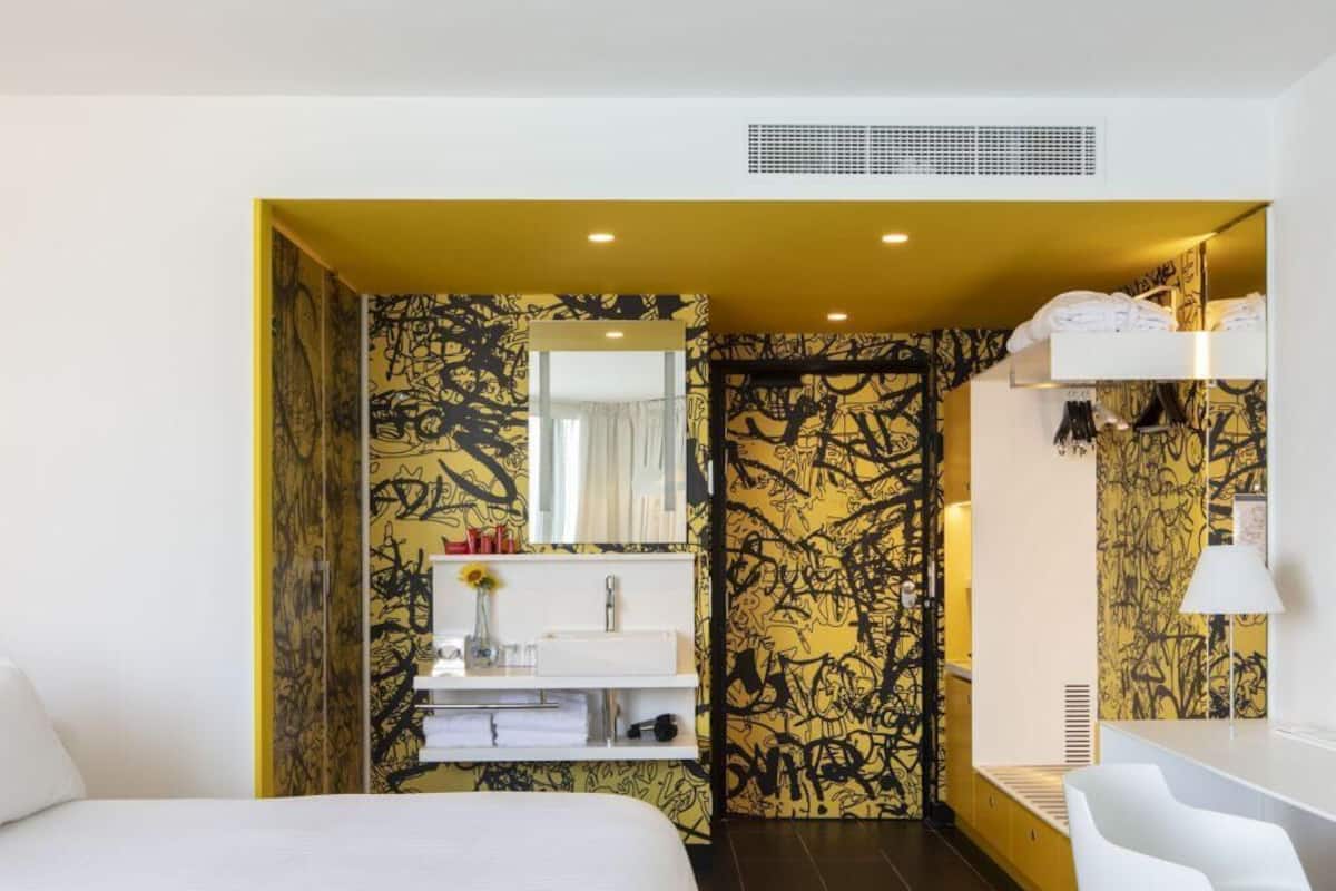 Habitación de hotel con cama, escritorio, baño con lavabo y espejo, y pared con diseño.