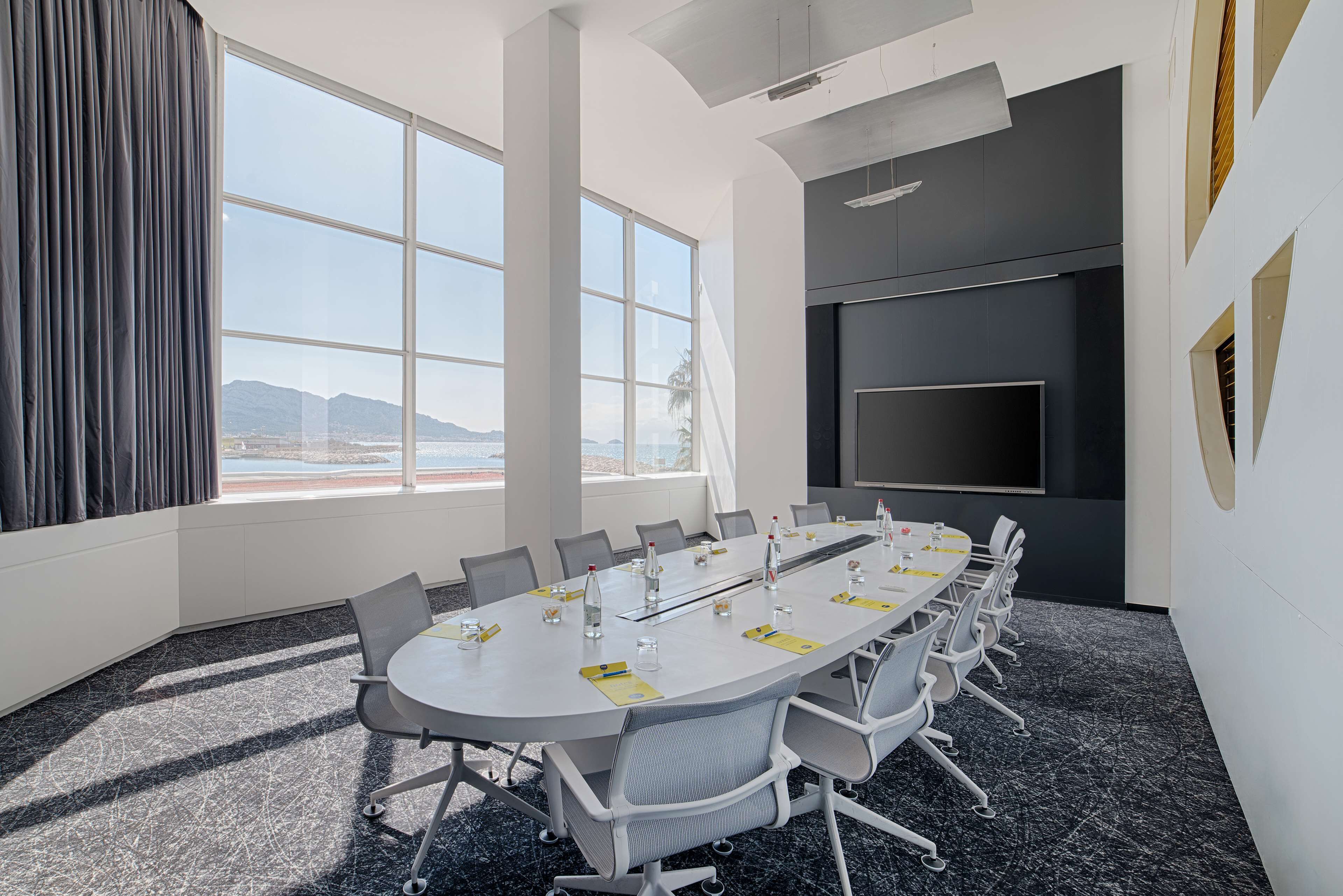 A conference room with a long table, chairs, a large window, and a flat-screen TV.